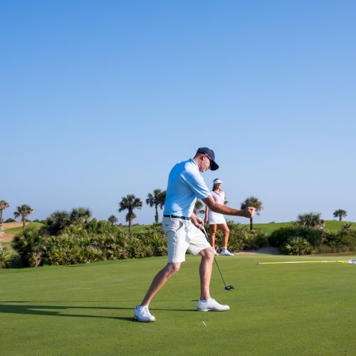 A man is playing golf on a sunny day, with another person and palm trees visible in the background on a well-maintained course.