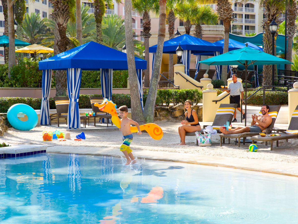 People enjoying a sunny day at a poolside with lounge chairs, umbrellas, and float toys, including a child about to jump into the pool.