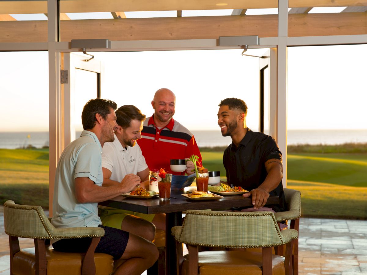 A group of four men sitting at a table enjoying a meal and drinks outdoors with a scenic view in the background.