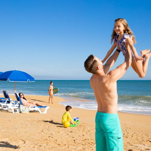 A man lifts a child playfully at a beach with umbrellas, sunbathers, and another child playing in the sand near the ocean.