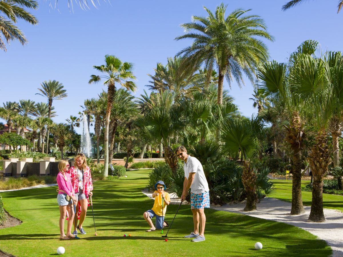 A family enjoys playing golf on a sunny day, surrounded by palm trees and lush greenery.