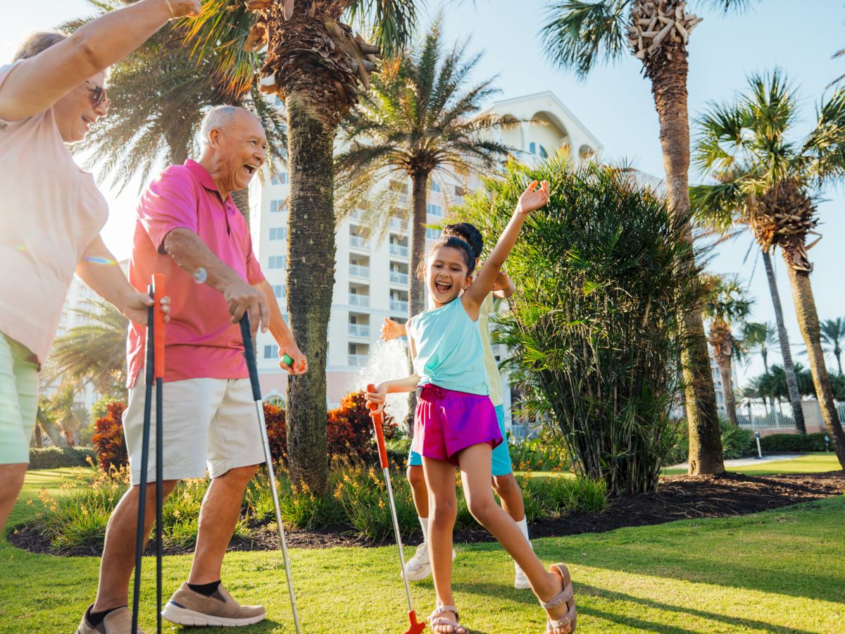 A joyful group of people playing a lawn game under sunny palm trees, with one girl celebrating.