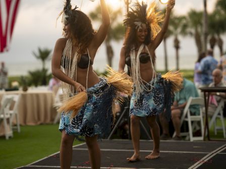 Two women perform a traditional dance wearing grass skirts and feathered headpieces on an outdoor stage.