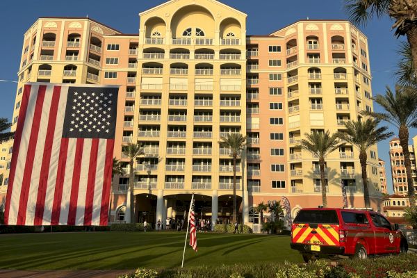 A large building displays a huge American flag, with a fire vehicle nearby and palm trees around, under a clear blue sky.