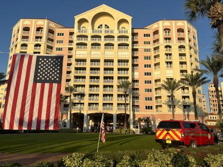 A large building with a massive American flag, a red vehicle with emergency markings, palm trees, and greenery in the foreground.