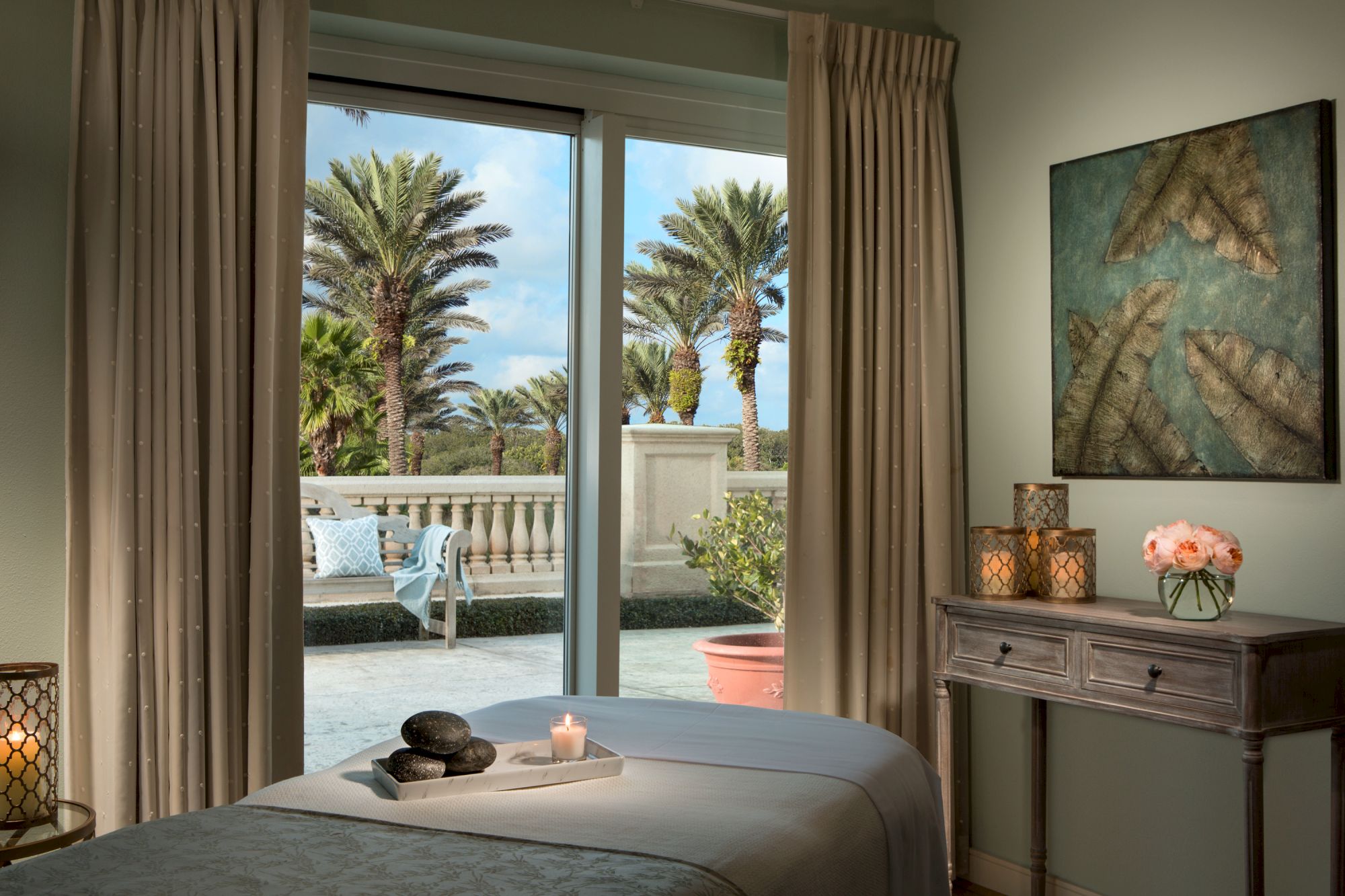 A serene room with a massage table, candles, and ocean view through a sliding door, leading to a terrace with palm trees.
