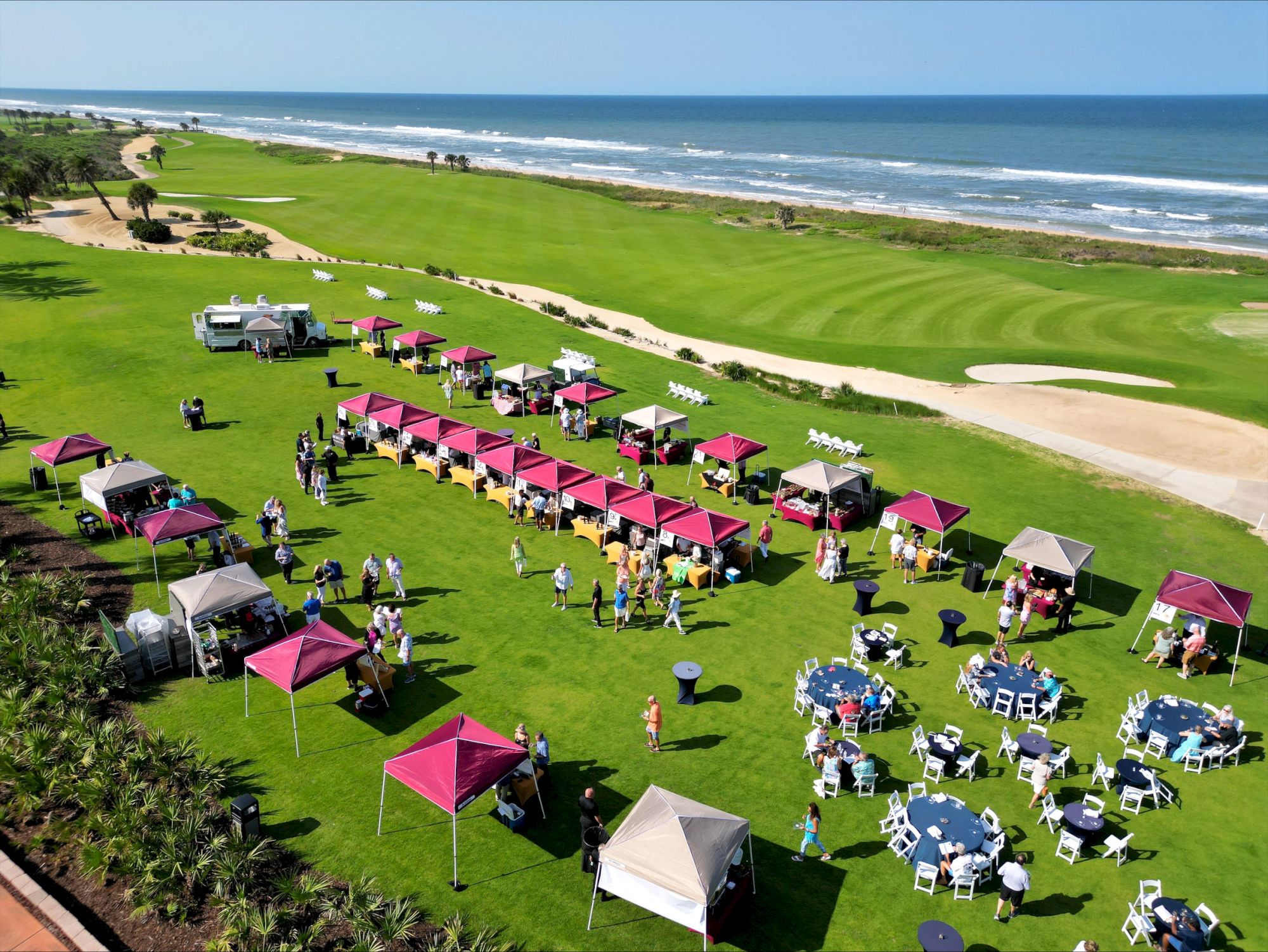 An outdoor event on a golf course by the ocean, with tents, tables, chairs, and people gathering under a clear blue sky.