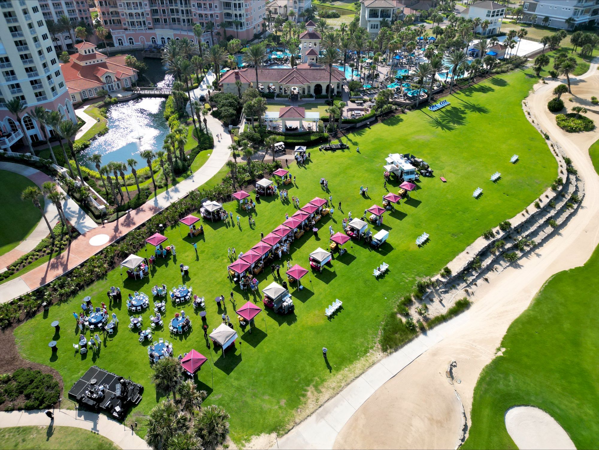 An aerial view of an outdoor event on a grassy area with tents, tables, and people, near pools and sand paths.