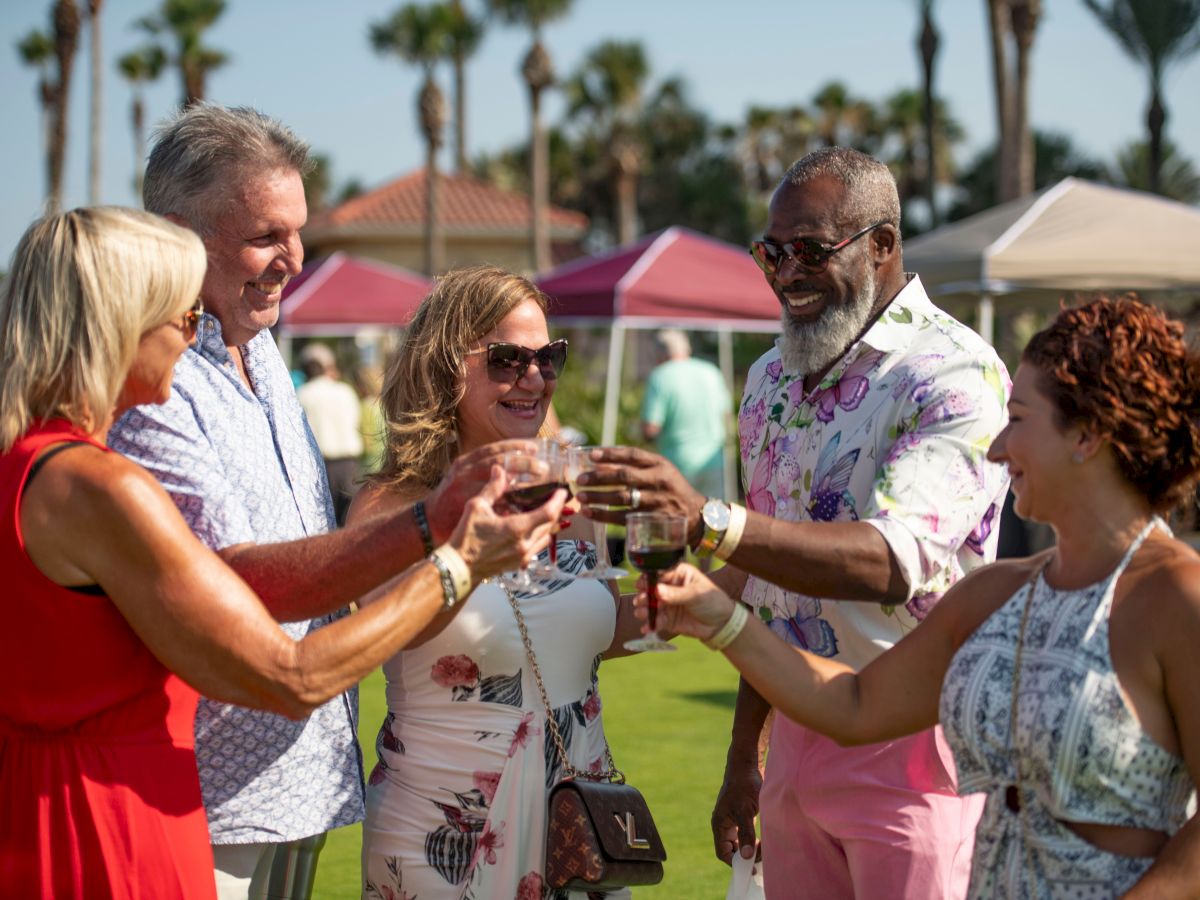 A group of five people outdoors toast with drinks. They're dressed in summer attire, enjoying a sunny day with palm trees in the background.