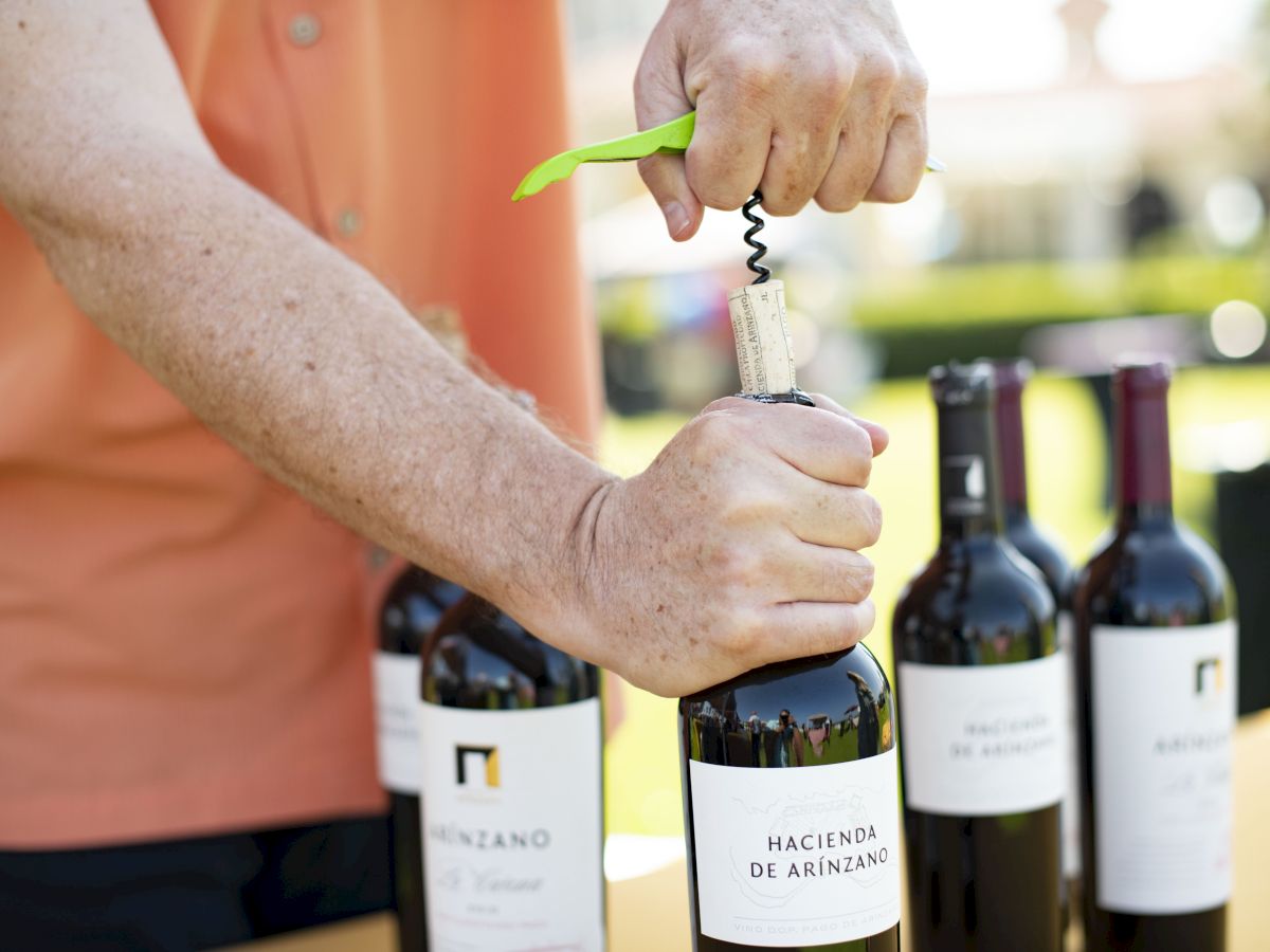 A person is opening a bottle of Hacienda De Arínzano wine with a corkscrew. Several other wine bottles are on the table.