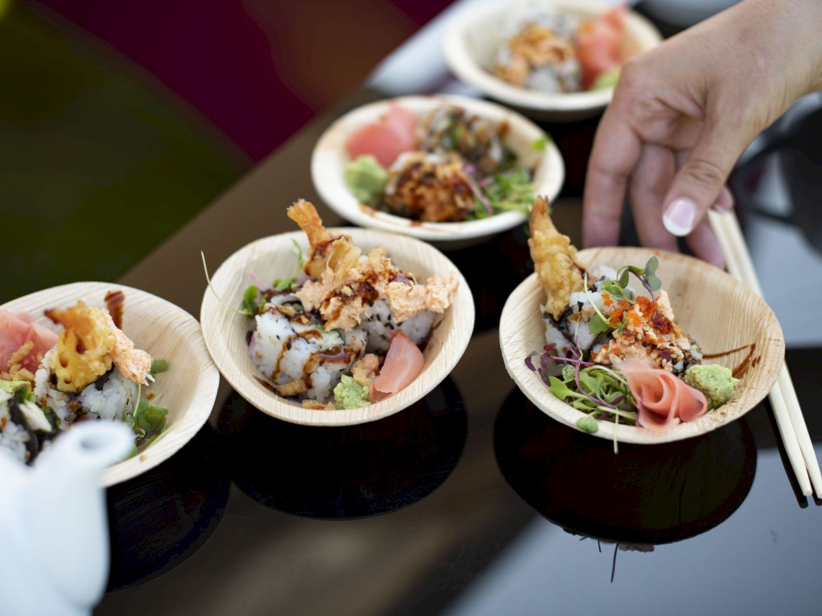 Small bowls of sushi with tempura, garnished with pickled ginger and wasabi, are being served with chopsticks.