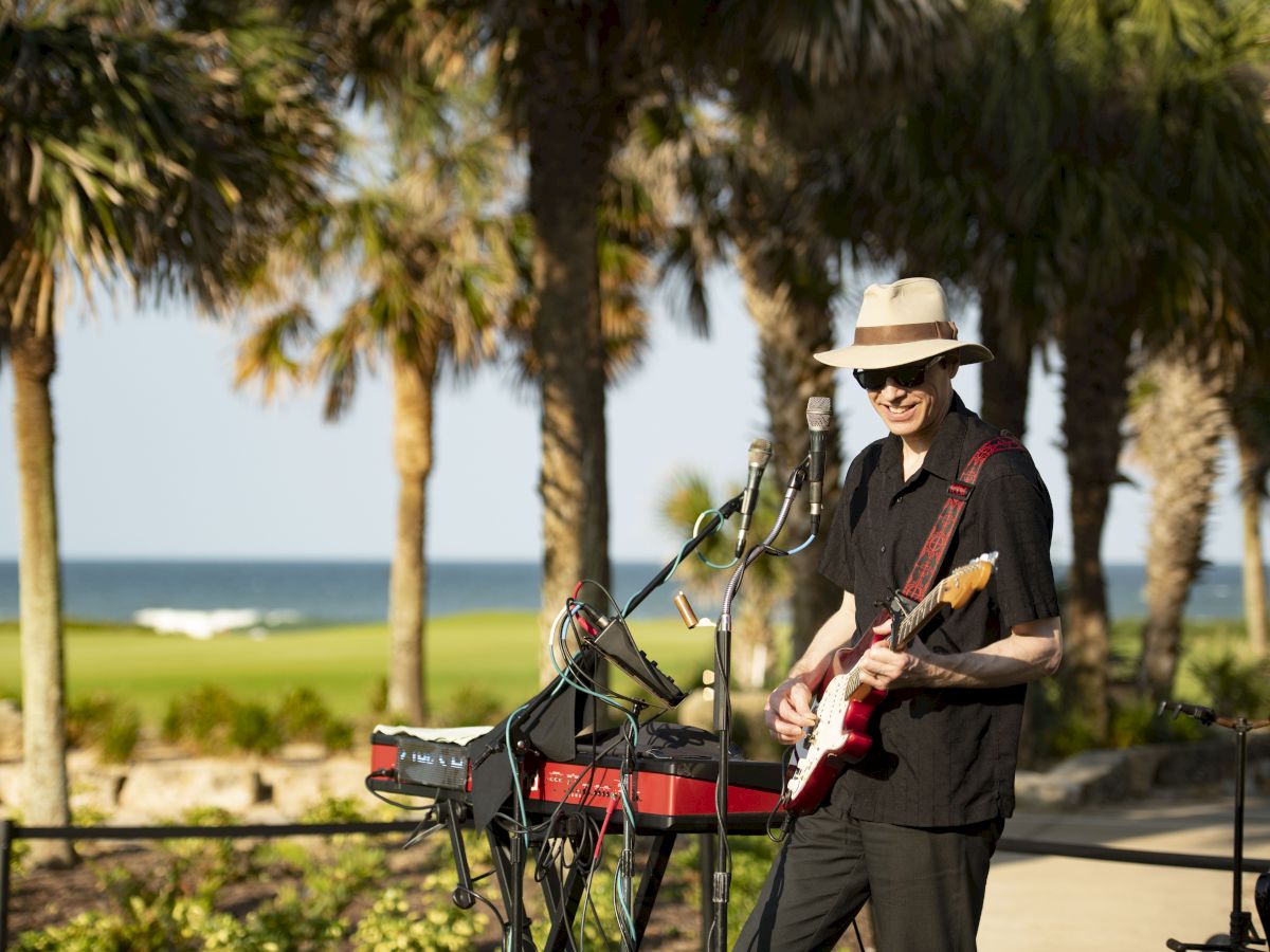 A musician in a hat plays guitar and keyboard outdoors, surrounded by palm trees, with the ocean in the background.