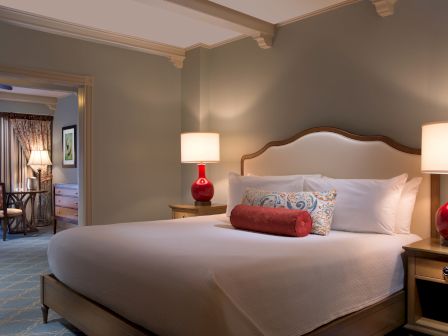 A cozy hotel room with a large bed, decorative pillows, bedside lamps, and an open door leading to another room with chairs and a table.