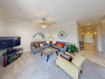 A cozy living room with a TV, sofas, coffee table, armchair, artwork, ceiling fan, plant, and decor in neutral tones. Bright hallway visible.