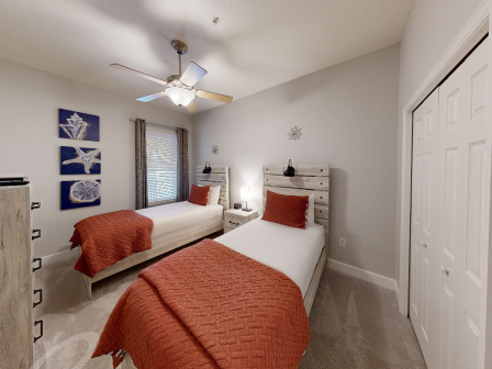 The image shows a bedroom with two twin beds, red accents, wall art, a ceiling fan, and a closet. The decor is bright and modern.