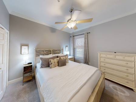 A cozy bedroom with a bed, decorative pillows, ceiling fan, dresser, and soft lighting. Neutral tones create a welcoming atmosphere.