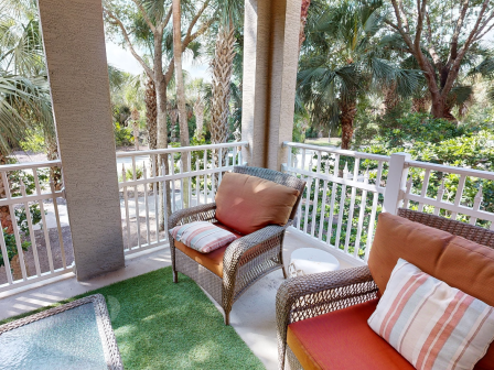 A cozy balcony with wicker chairs and cushions overlooks lush green trees, creating a relaxing outdoor space.