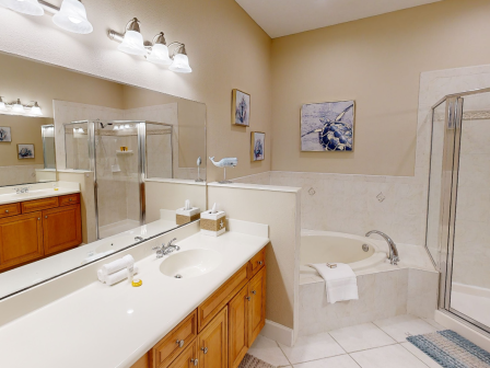 The image shows a modern bathroom with a large mirror, double sink, wooden cabinets, a bathtub, shower, and artwork on beige walls.