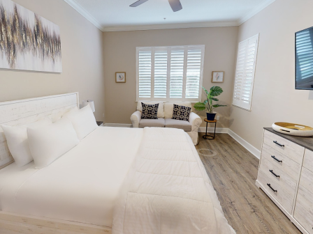 A neatly arranged bedroom with a white bed, sofa, decor on walls, a TV, and a dresser with a decorative tray on it.
