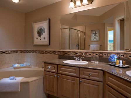 A bathroom with a tub, double sink vanity, wall art, and a mirror. Neutral tones with decorative tiles and lighting above the mirror.