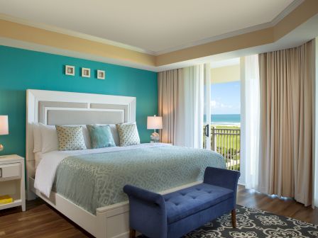 A well-furnished bedroom with a large bed, blue bench, teal wall, lamps, and a balcony view of the ocean through sheer curtains.