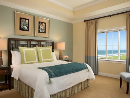 A coastal-themed bedroom features a bed with green accents, art above, side tables with lamps, and a window showing a beach view.