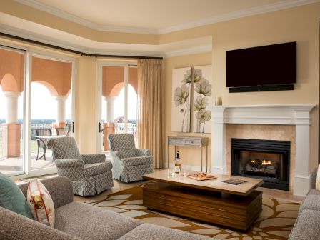 A cozy living room features a fireplace, TV, sofa, armchairs, coffee table with wine, and a balcony view with archways.