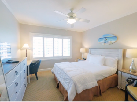 A cozy bedroom with a neatly made bed, two bedside lamps, a dresser, wall art, and a window with shutters under a ceiling fan.