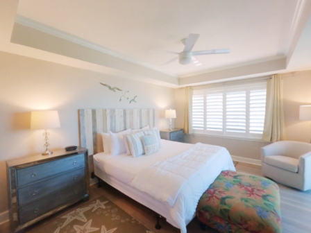A cozy bedroom with a bed, armchair, two nightstands, and lamps, featuring a ceiling fan and a window with shutters.
