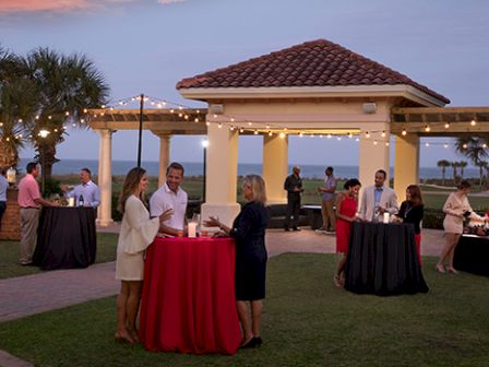 An outdoor gathering at a pavilion near the beach, with people chatting and standing around tables adorned with red tablecloths under string lights.