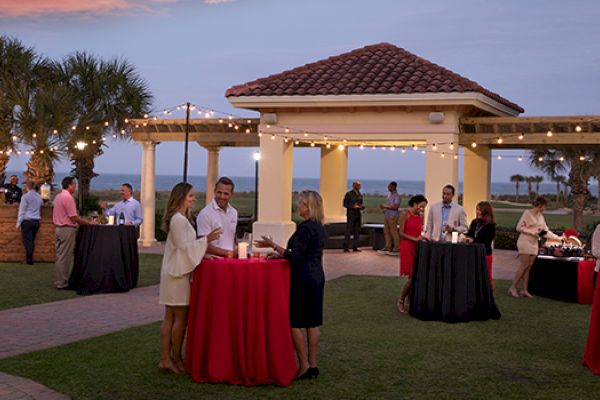 An outdoor gathering at a pavilion near the beach, with people chatting and standing around tables adorned with red tablecloths under string lights.