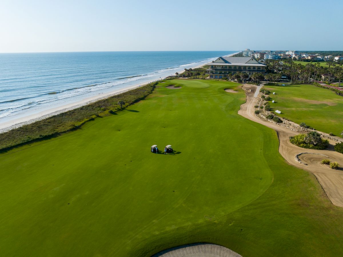 A golf course by the ocean, with two golf carts on the green, surrounded by sand dunes and buildings in the distance.