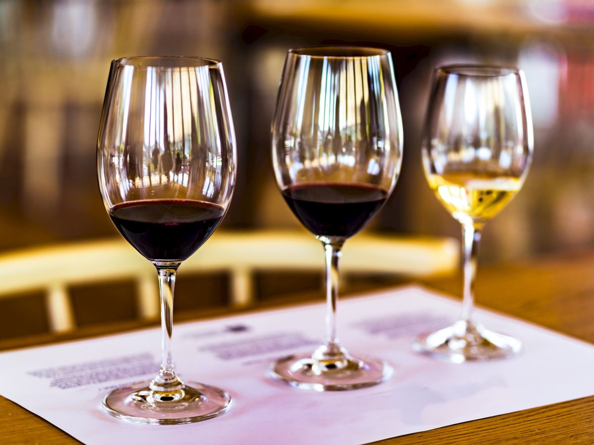 Three wine glasses are on a table. Two are filled with red wine, and one with white wine, placed on a paper with text.