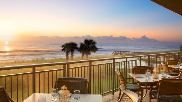 A seaside dining area on a balcony with set tables overlooks the ocean at sunrise, with a peaceful and scenic view beyond the railing.