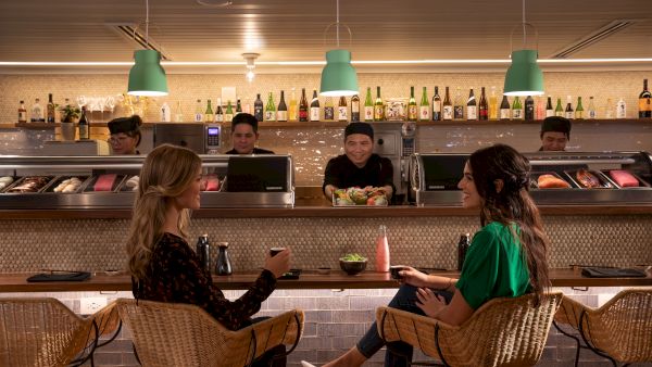 Two people sit at a sushi bar, chatting and having drinks. Chefs behind the counter prepare sushi. Bottles line the background shelf.