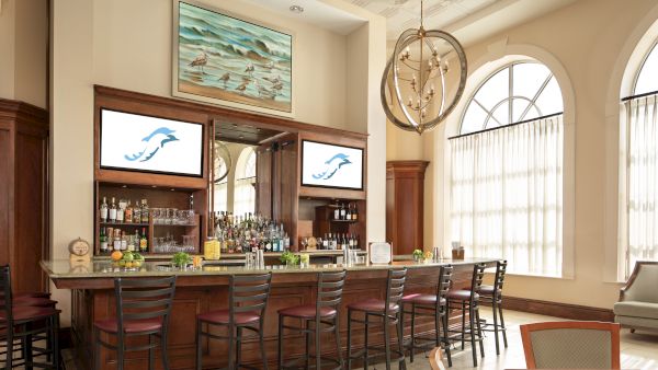 A stylish bar with high chairs, bottles, and decor. There are screens above and large windows in the background.