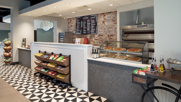 This image shows a modern pizza shop interior with a patterned floor, snack display, pizza counter, and menu on the wall.