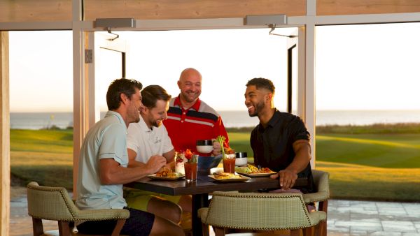 A group of four men are laughing and enjoying a meal together in a bright room with an outdoor view.