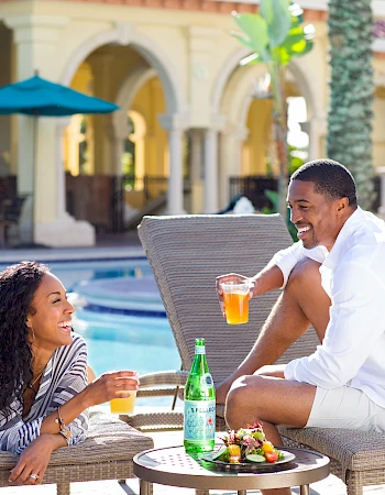 A couple relaxes by a poolside with drinks and snacks, enjoying the sun and each other's company at a resort-like setting.