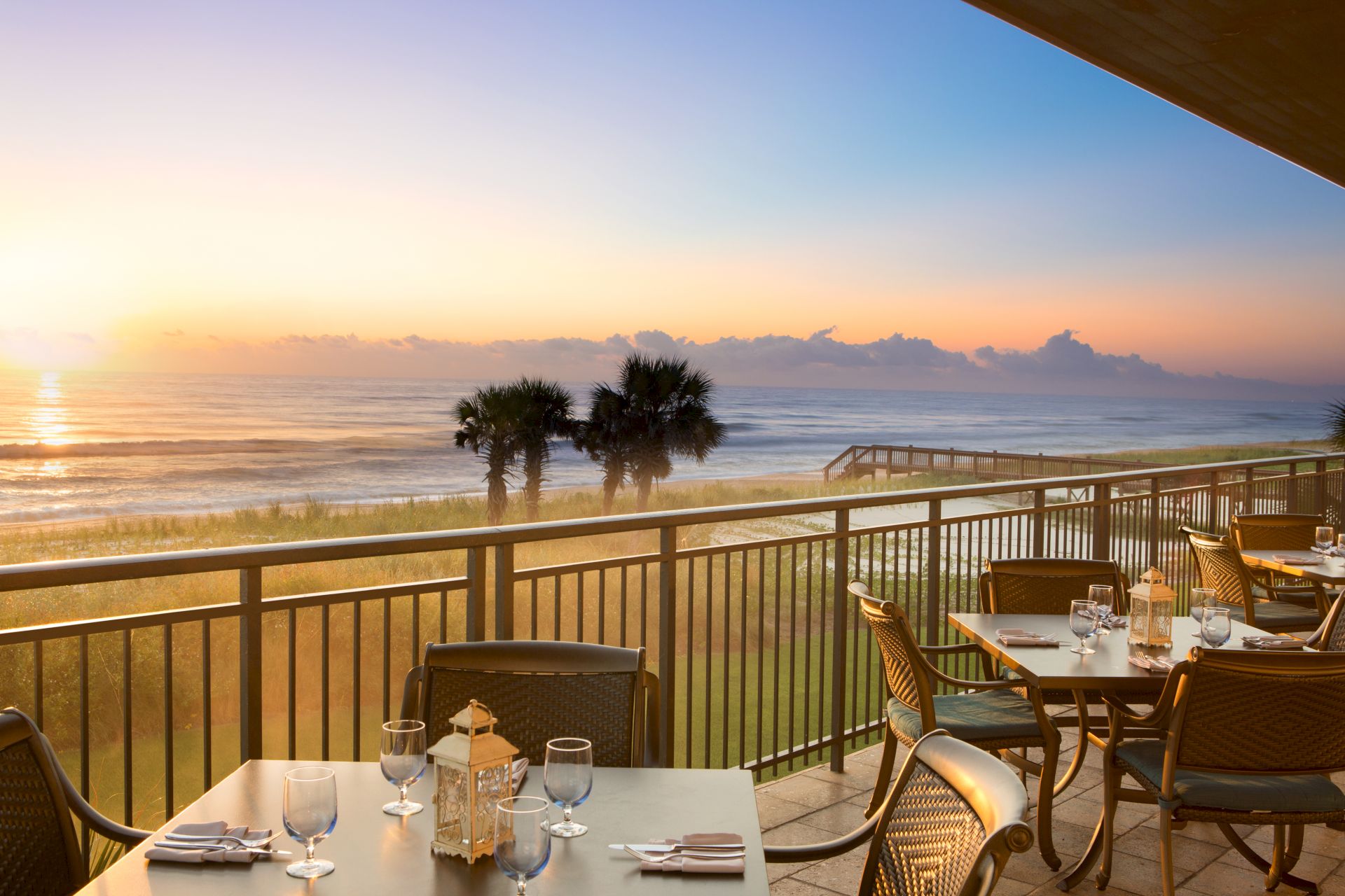 A beachside dining area with set tables overlooks the ocean. The sun is setting over the water, creating a peaceful ambiance.