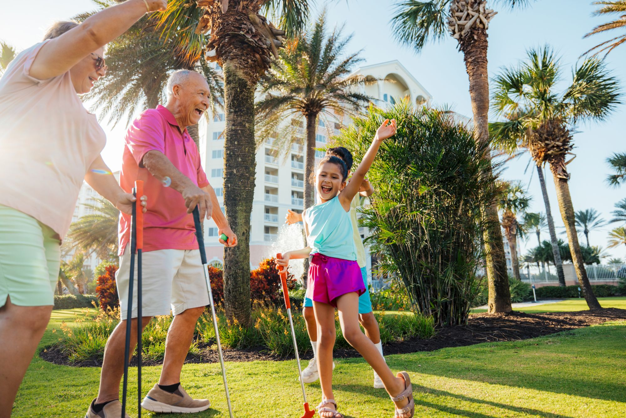 A joyful group plays mini-golf on a sunny day, surrounded by palm trees, with a girl celebrating a putt and others cheering.