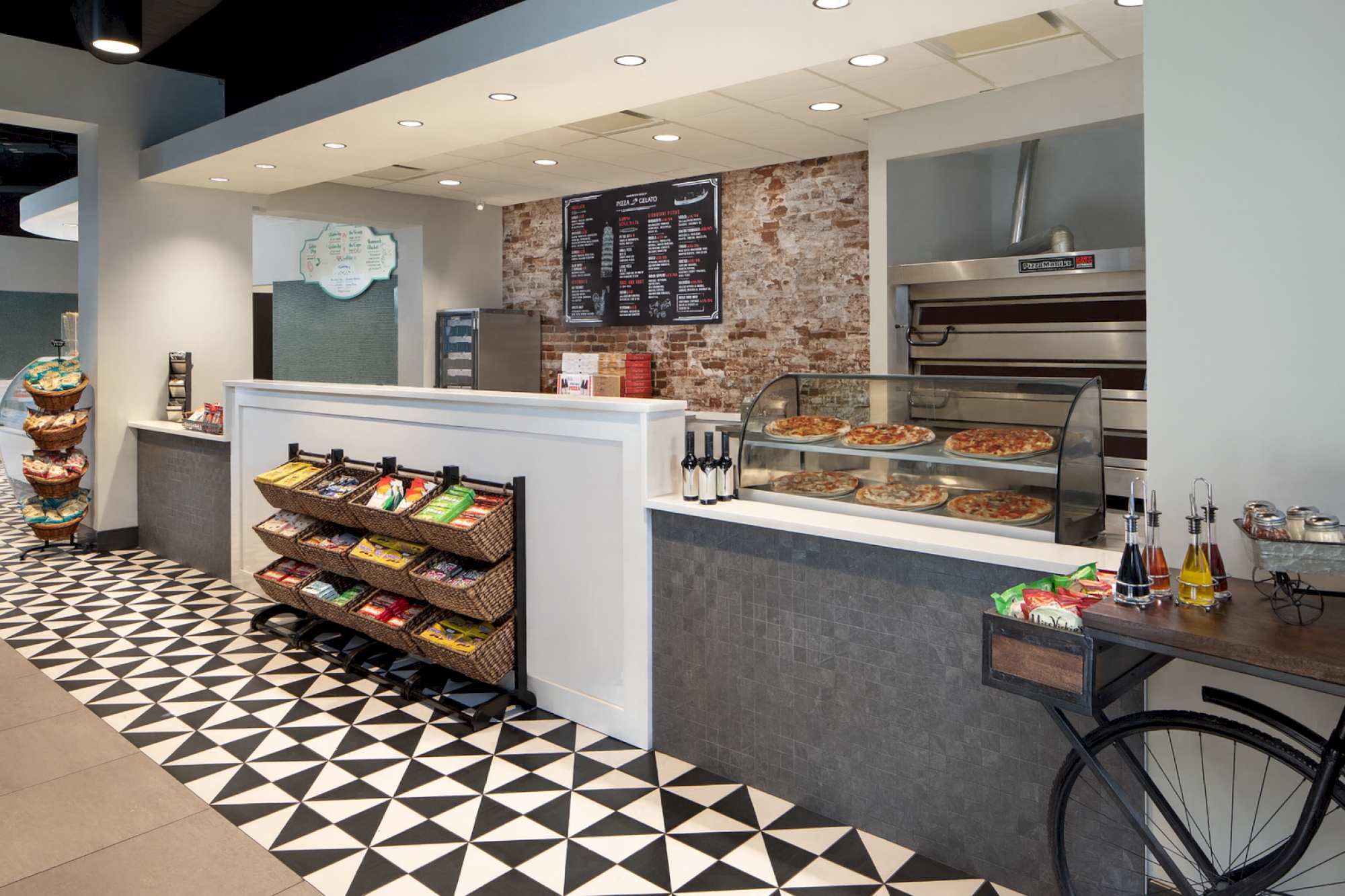 The image shows a modern pizzeria interior with a counter, pizza display, snacks, drinks, and patterned floor tiles.
