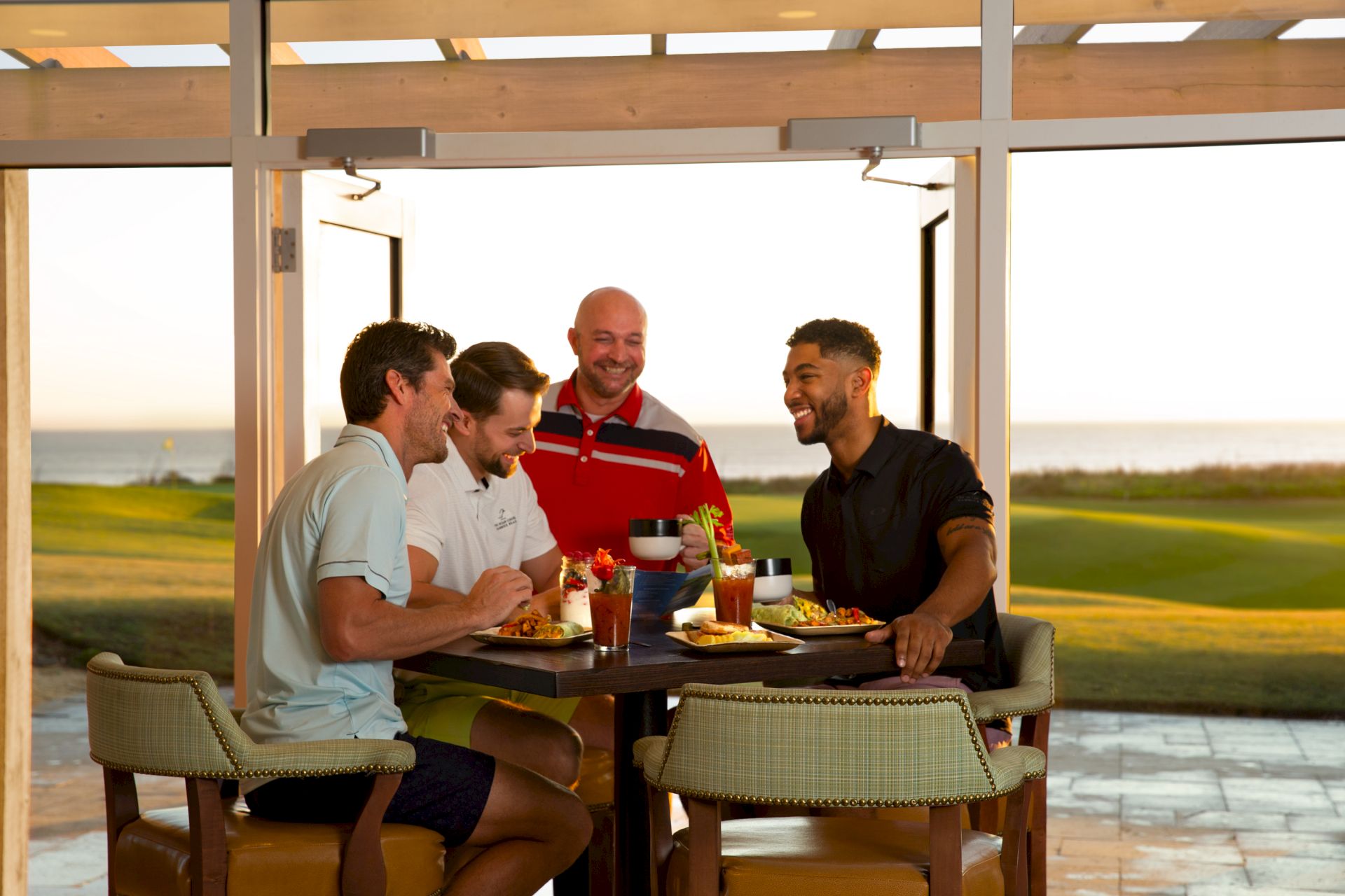 A group of four men are seated at a table outdoors, enjoying food and drinks with a scenic view in the background.