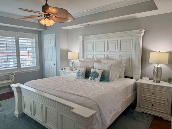 A cozy bedroom features a large bed with white bedding, complemented by matching white nightstands, lamps, and a ceiling fan.