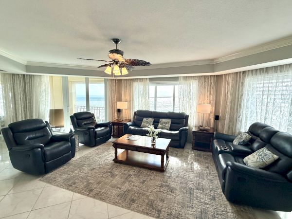 The image shows a living room with black leather sofas, a coffee table, and lamps, set in front of large windows with sheer curtains.