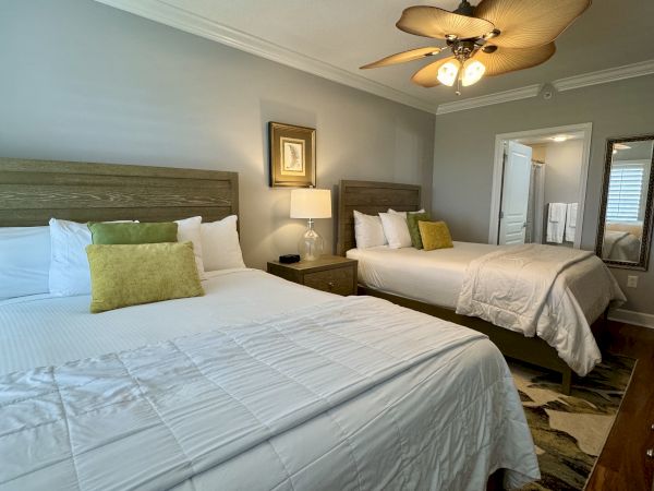 A cozy bedroom with two beds, a side table, lamp, ceiling fan, and artwork. Light-colored walls and a door leading to a bathroom are visible.