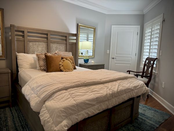 A cozy bedroom with a wooden bed, neutral bedding, decorative pillows, a side table with a lamp, a mirror, and a chair by the window.