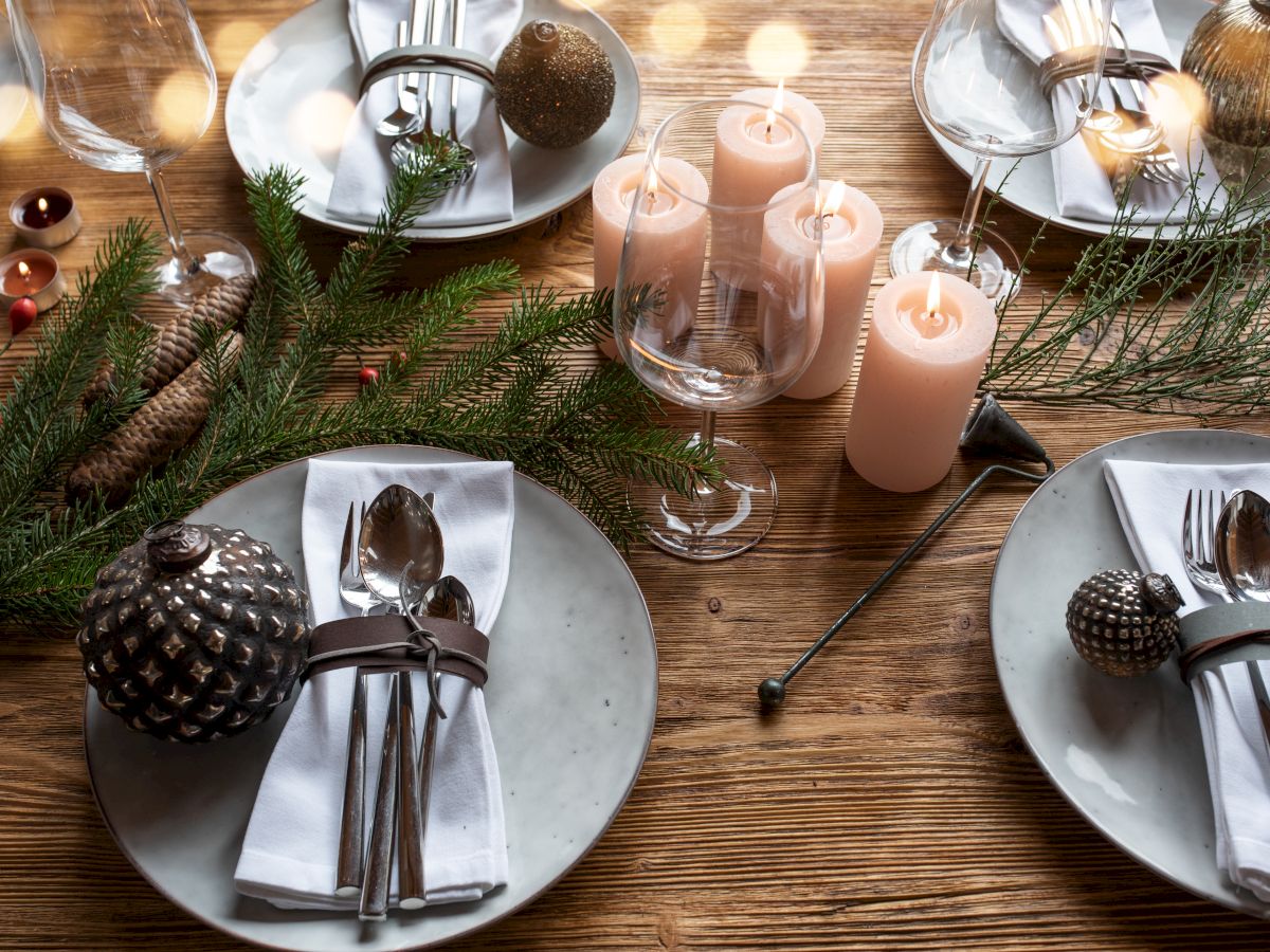 A festive holiday table set with lit pink candles, pine branches, pinecones, and glasses and plates for a cozy dinner party.