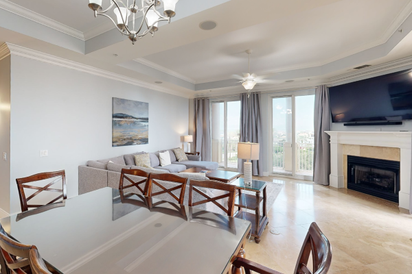 A modern living room with a gray sofa, dining table, large TV, fireplace, chandelier, artwork, and glass doors leading to a balcony.