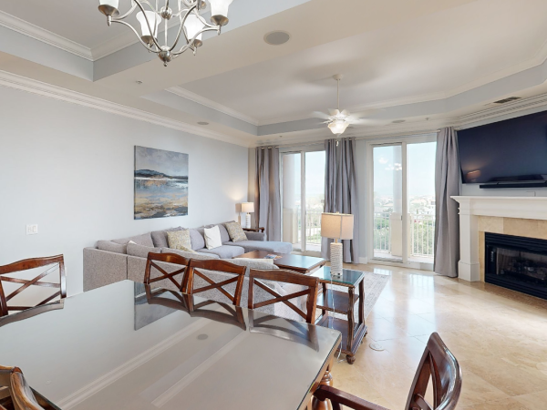 A modern living room with a gray sofa, dining table, large TV, fireplace, chandelier, artwork, and glass doors leading to a balcony.